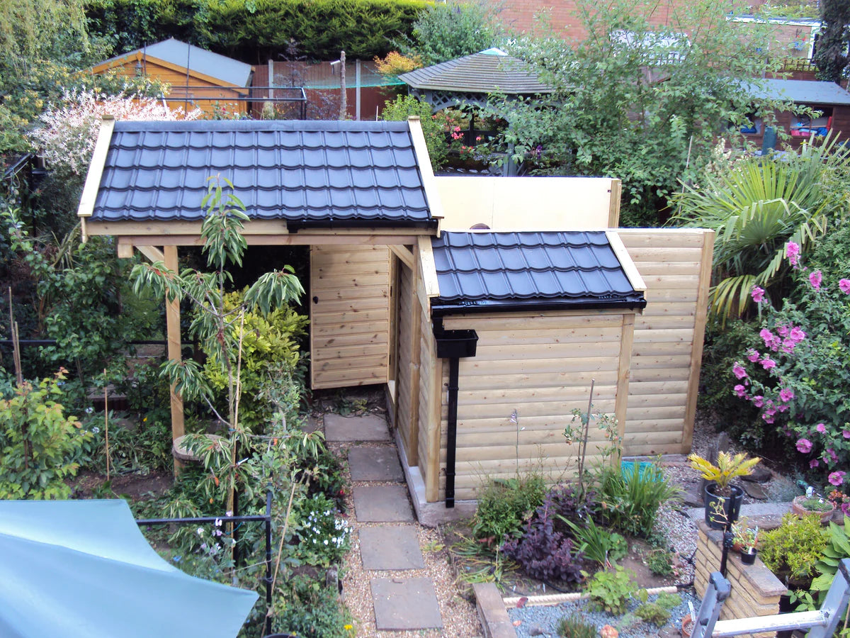 Smooth black lightweight roof tiles on a small, two-part wooden garden observatory building, with the two sections of the roof slightly separated. The building is surrounded by a lush garden.
