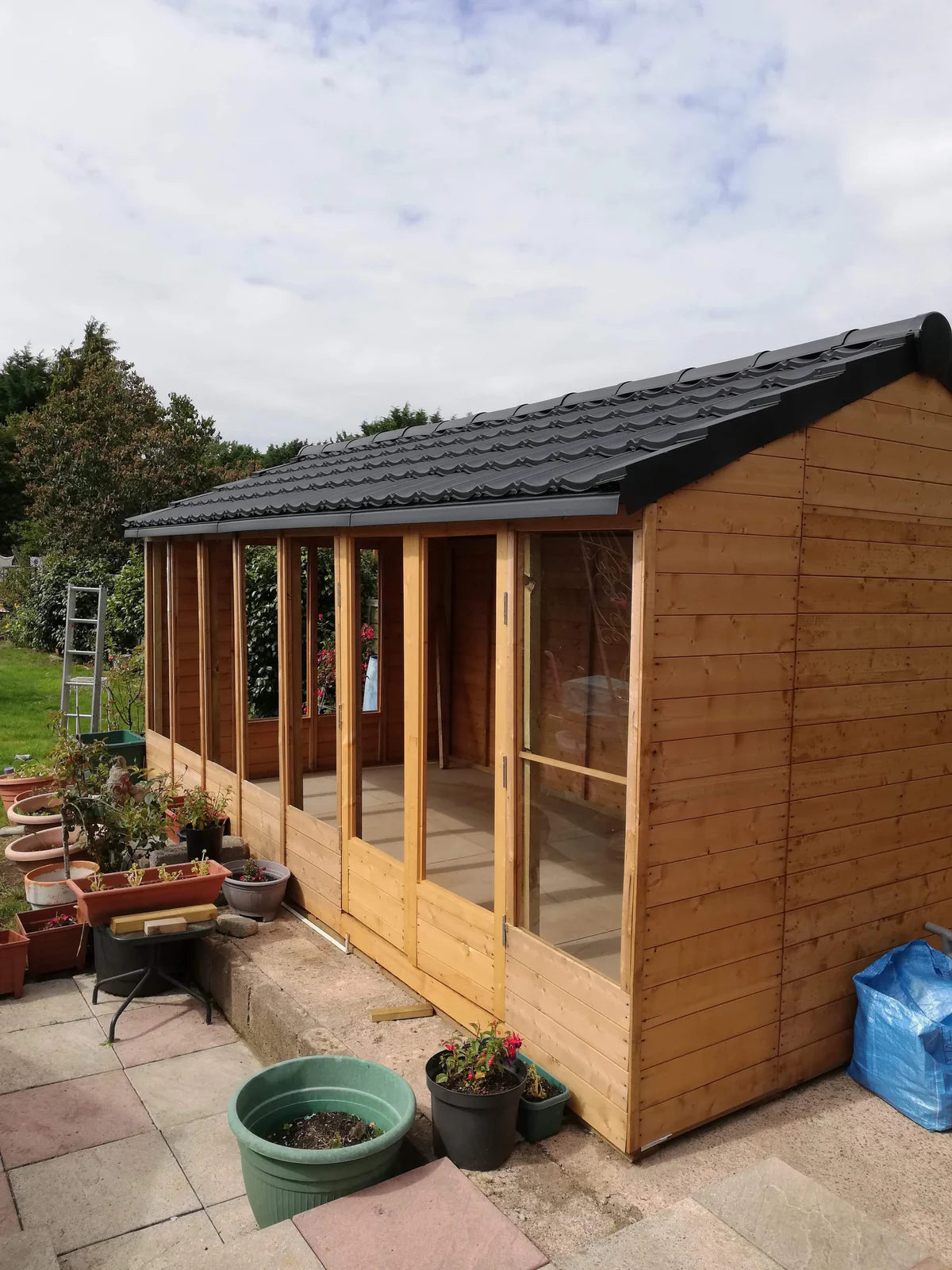 A side view of a wooden summerhouse with black lightweight roof tiles. The building features large windows and is set on a paved patio with various potted plants.