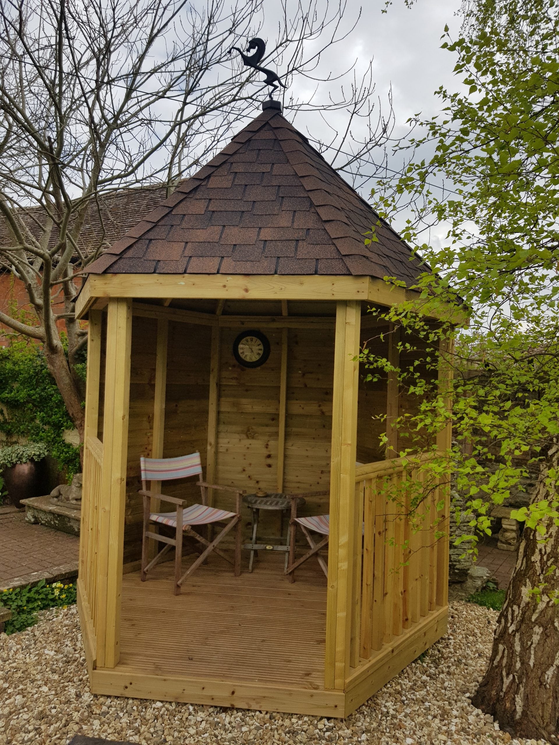 Wooden gazebo with an ochre roof, chairs, and a clock in a garden setting.