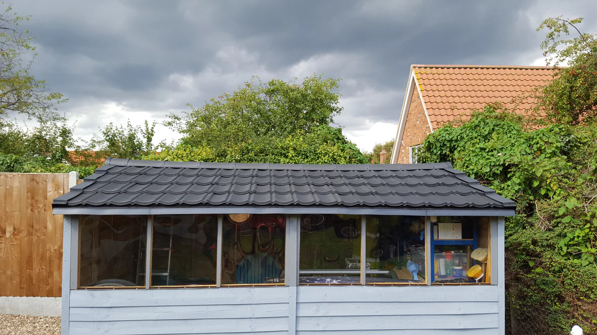 A long, horizontal garden shed with a black lightweight tiled roof. The shed is painted a light blue or gray and has a large, clear plastic window across the front.